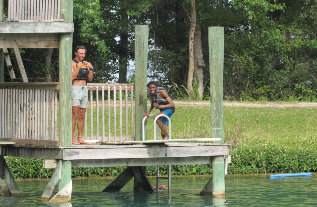 Two young people stand on a dock near a body of water with life jackets on during a summer camp.