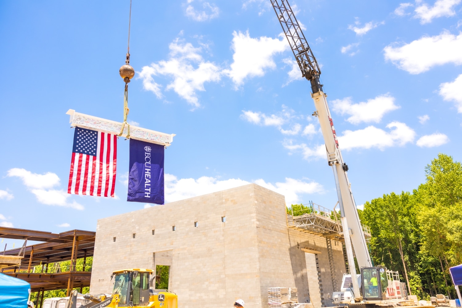 A beam, signed by ECU Health and Thomas Construction team members, is raised into position at the future site of a behavioral health hospital in Greenville. A United States of America Flag and ECU Health flag hang from the beam.